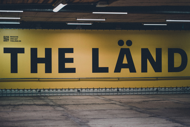 Ein großes gelbes Banner mit schwarzer Schrift ist über die gesamte Breite und Höhe einer Wand gespannt, darauf steht „The Länd“. Foto von Marcel Strauß via unsplash.com: https://unsplash.com/photos/a-yellow-sign-that-says-the-land-on-it-1bFTFTGUlt0